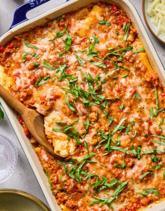 Baked pasta casserole topped with melted cheese and chopped fresh basil, with a wooden spoon serving a portion from a rectangular dish.