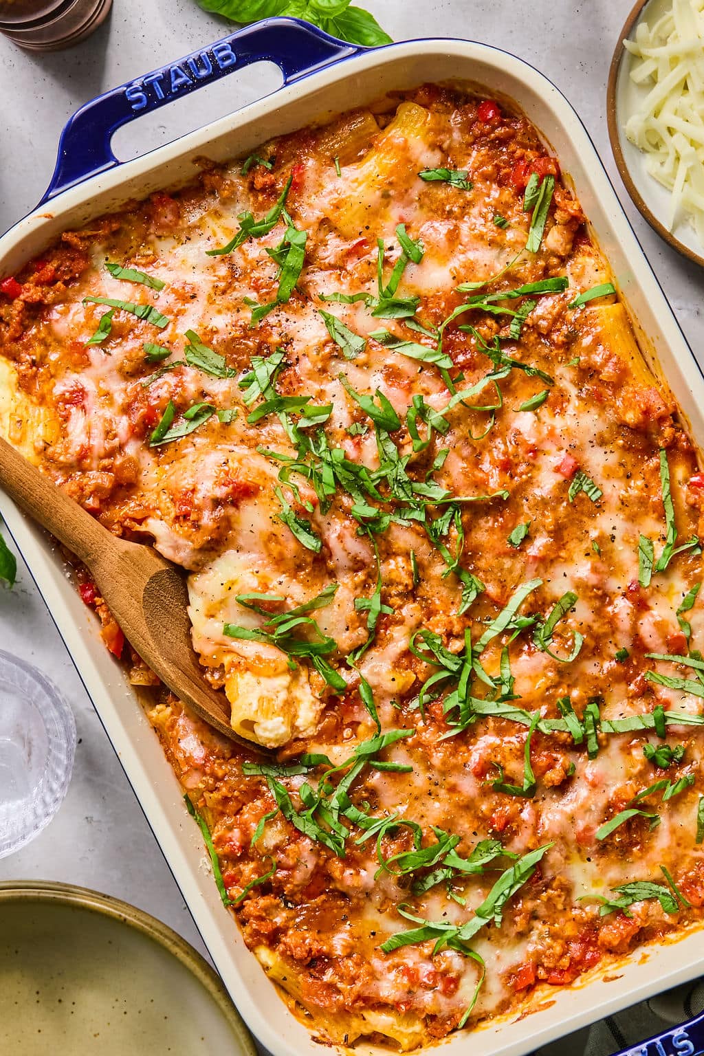 Baked pasta casserole topped with melted cheese and chopped fresh basil, with a wooden spoon serving a portion from a rectangular dish.