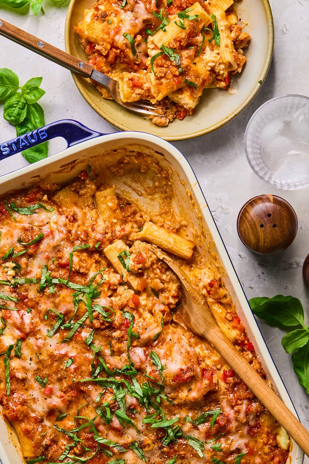 Baked pasta in a casserole dish topped with melted cheese and herbs, with a wooden spoon and a portion served on a plate nearby.