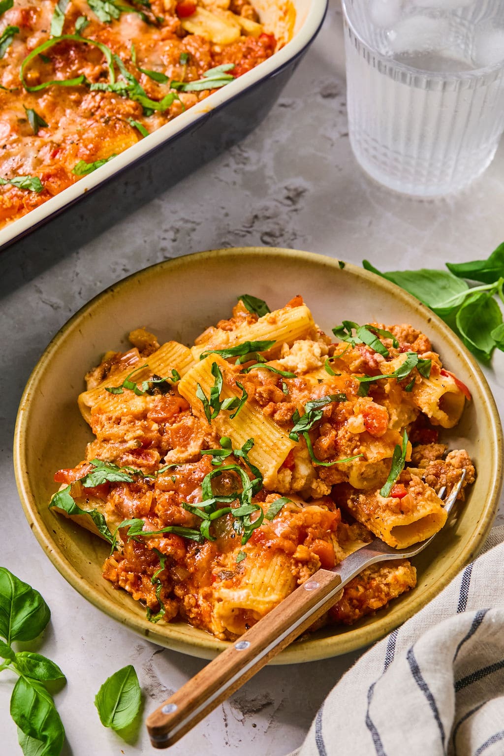 Bowl of baked rigatoni pasta with tomato sauce, cheese, and fresh basil, served with a fork. Dish and glass of water in the background.