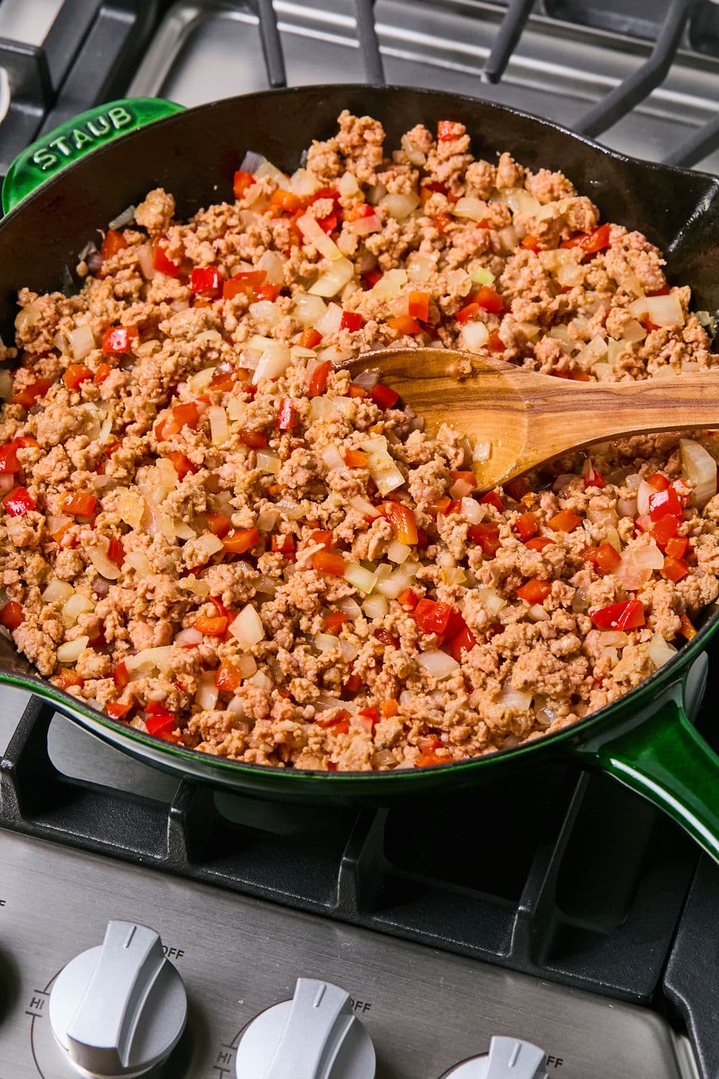 Ground meat, diced onions, and red bell peppers cooking in a green skillet on a stove, stirred with a wooden spoon.