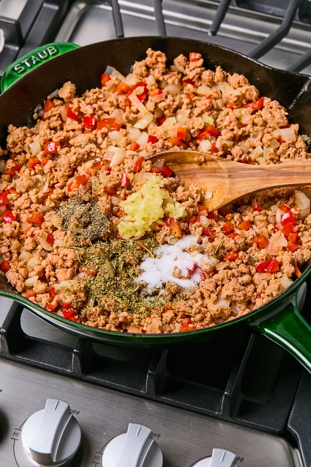 Ground meat, diced onions, and red bell peppers cooking in a skillet, with piles of spices, salt, and minced garlic on top; a wooden spoon rests in the pan.