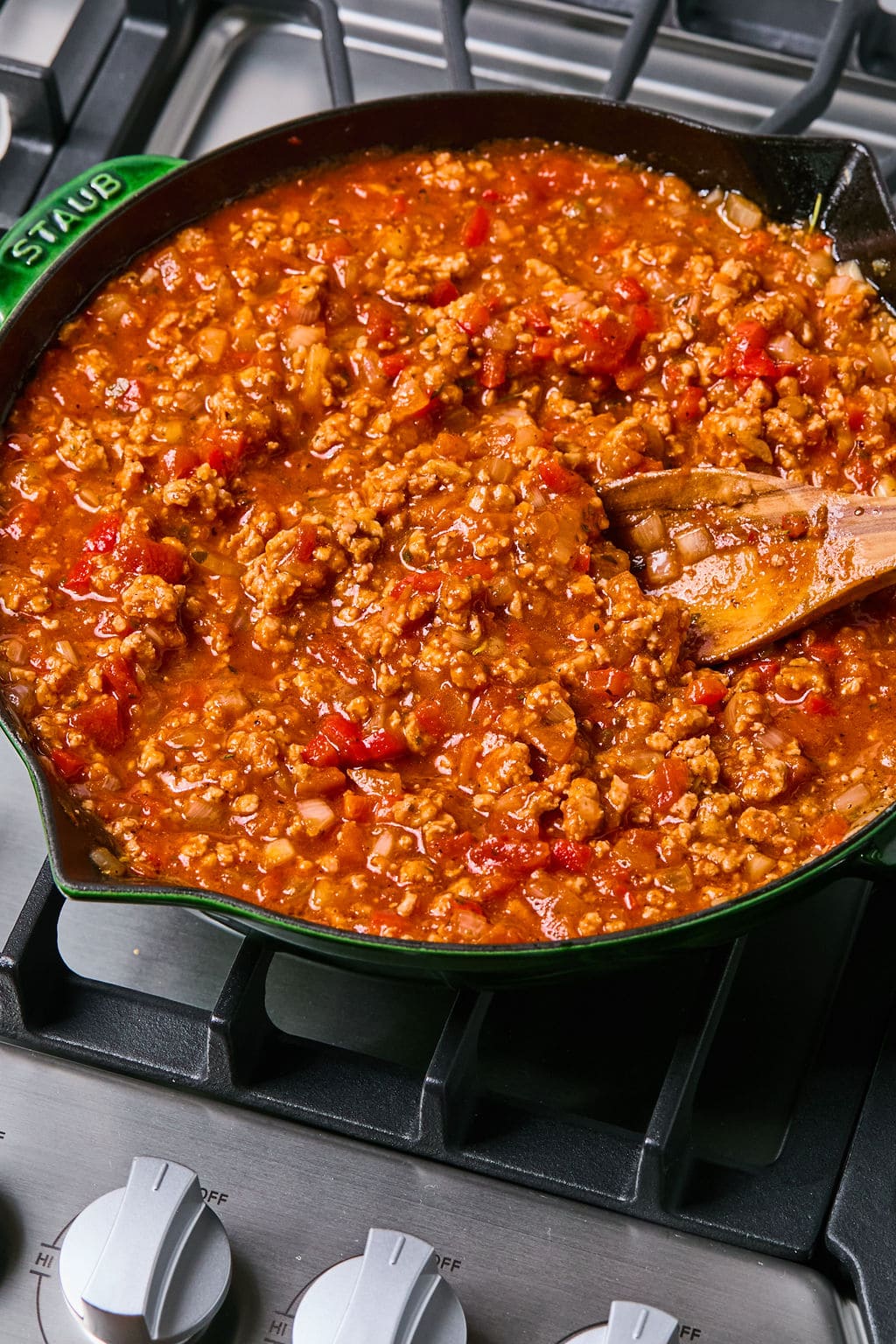 A skillet filled with ground meat, tomatoes, and sauce simmers on a stovetop, with a wooden spoon stirring the mixture.