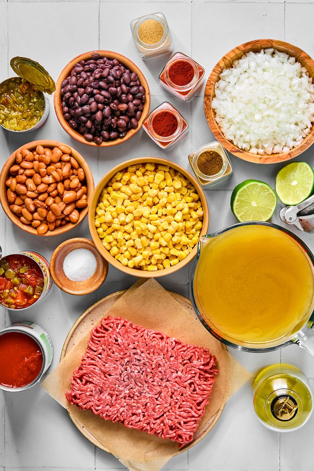 Raw ground beef, corn, black beans, pinto beans, chopped onion, lime halves, broth, olive oil, spices, salt, diced tomatoes, and sliced green chilies arranged on a white surface.