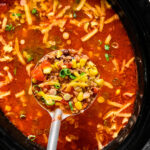 A hand holds a ladle of soup with ground beef, corn, tomatoes, beans, and green onions over a pot filled with the same soup, topped with shredded cheese.