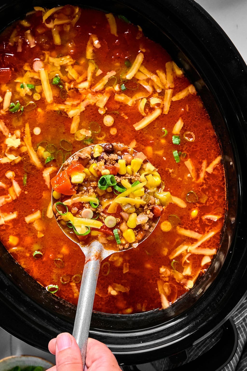 A hand holds a ladle of soup with ground beef, corn, tomatoes, beans, and green onions over a pot filled with the same soup, topped with shredded cheese.