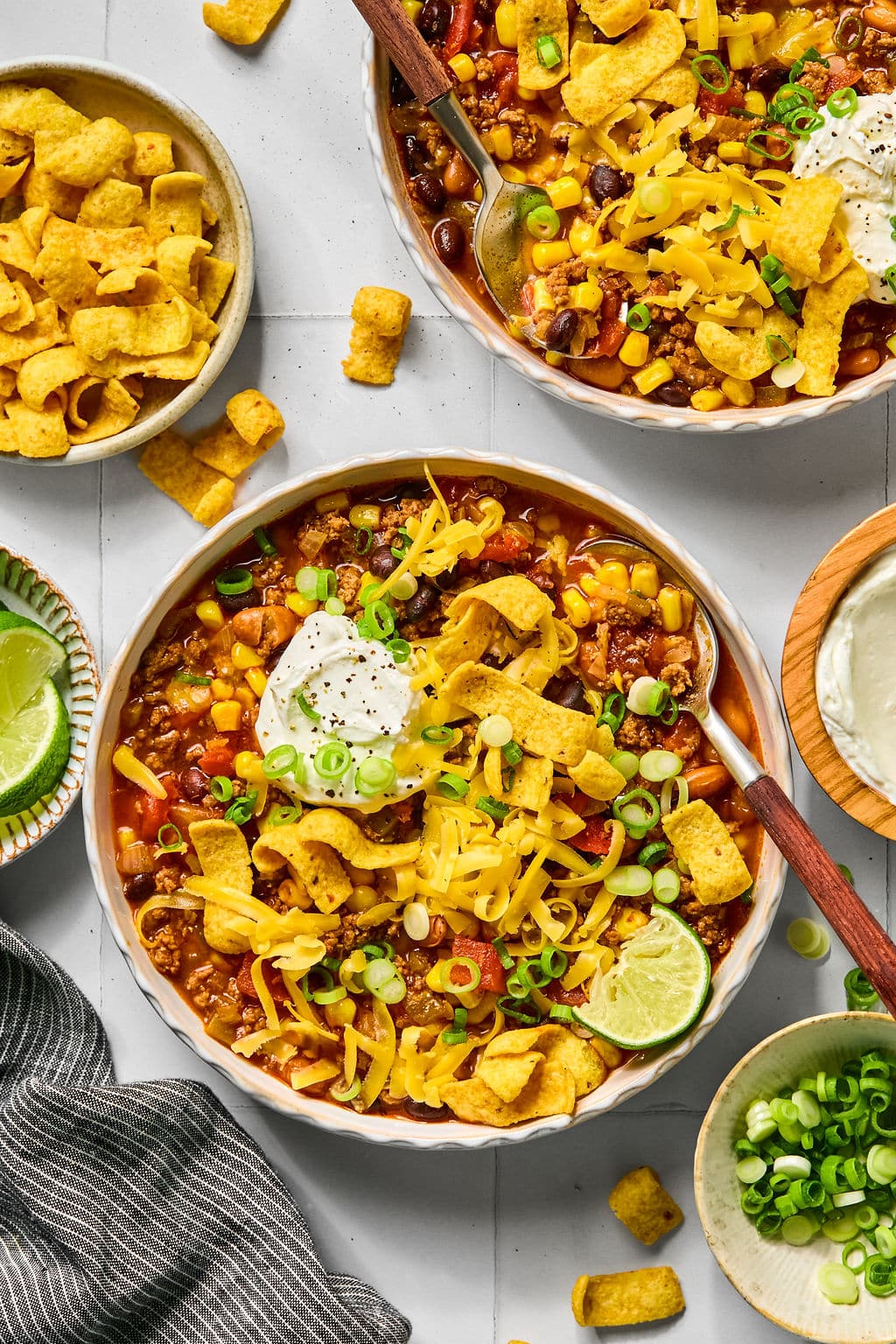 Two bowls of chili topped with shredded cheese, sour cream, corn chips, green onions, and lime wedges, surrounded by additional toppings and a striped napkin.