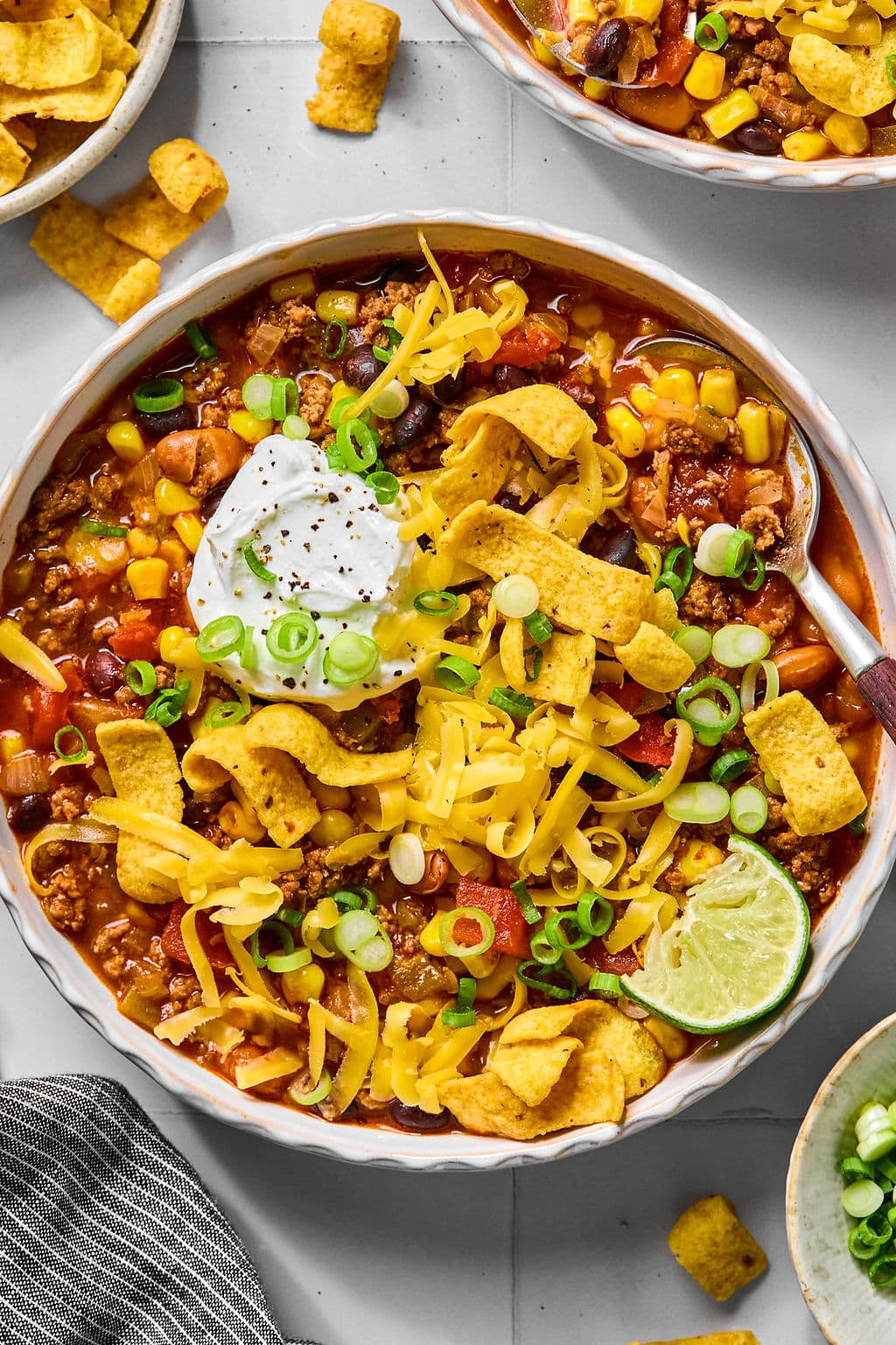 A bowl of chili topped with shredded cheese, corn chips, sour cream, sliced green onions, and a lime wedge, with a spoon on the side.