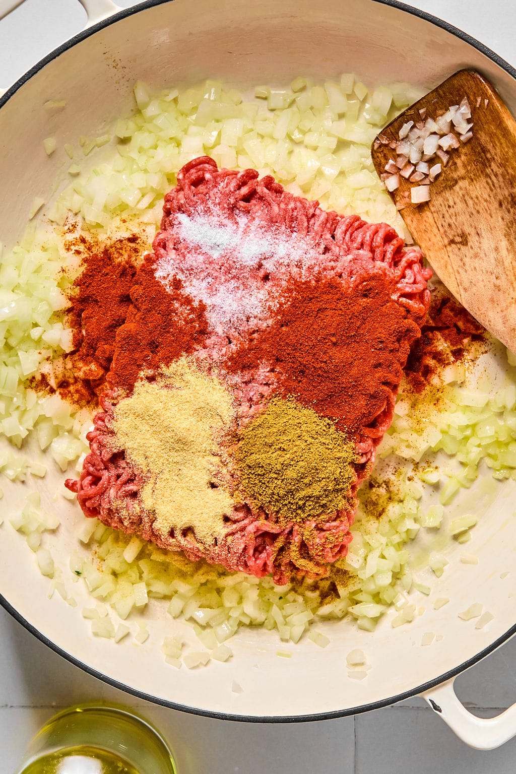 Ground beef with spices and chopped onions cooking in a large white pan, with a wooden spatula resting on the side.