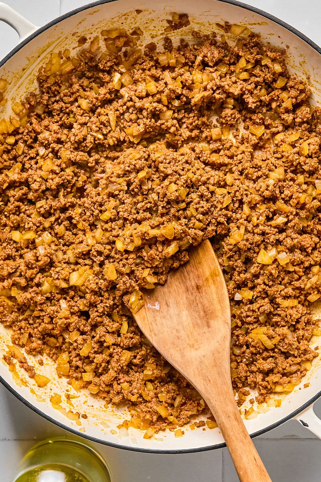 Ground beef and diced onions cooking in a white skillet with a wooden spatula, seen from above.
