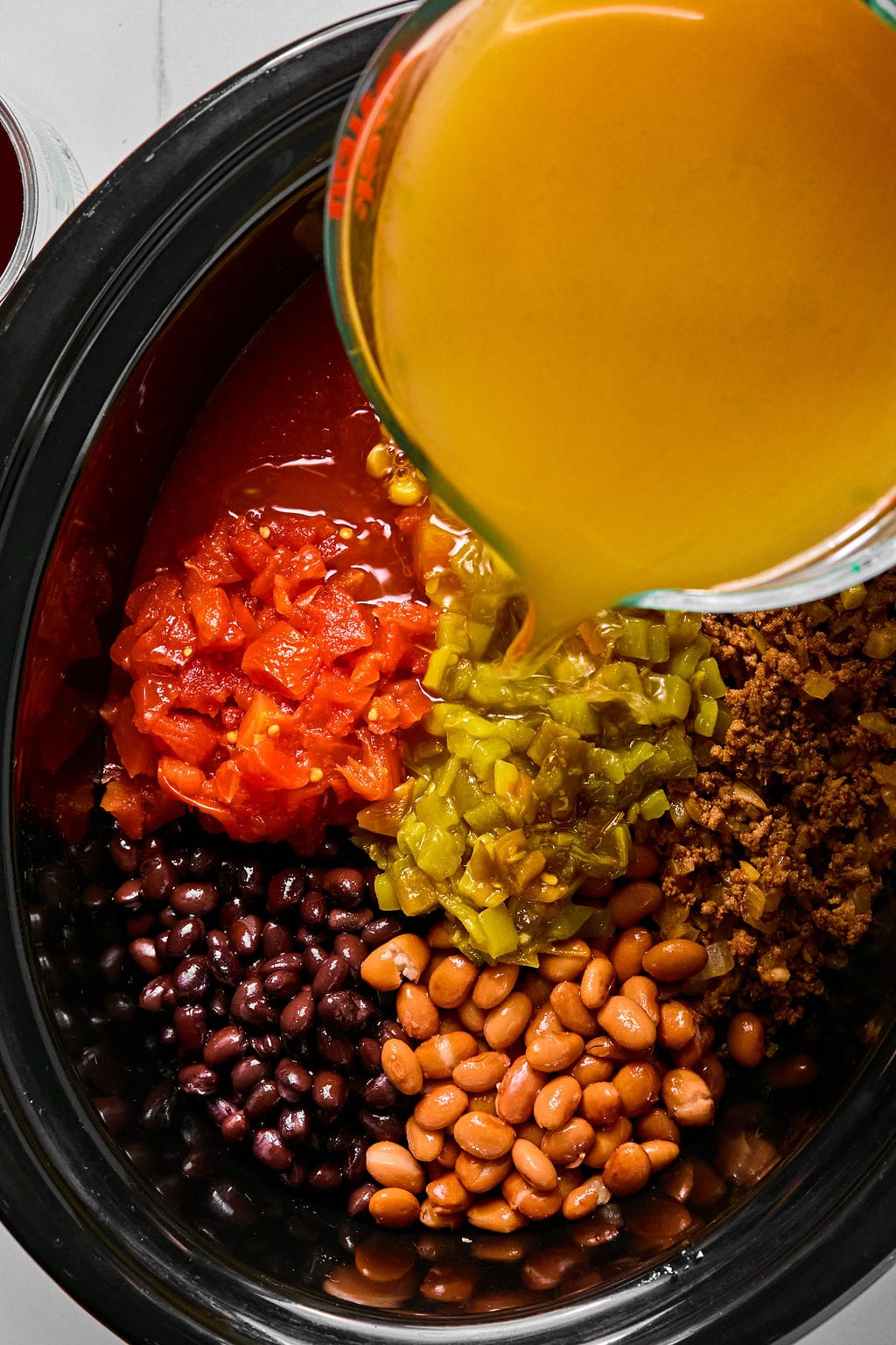 Overhead view of beans, tomatoes, green chilies, ground meat, and broth being poured into a slow cooker.
