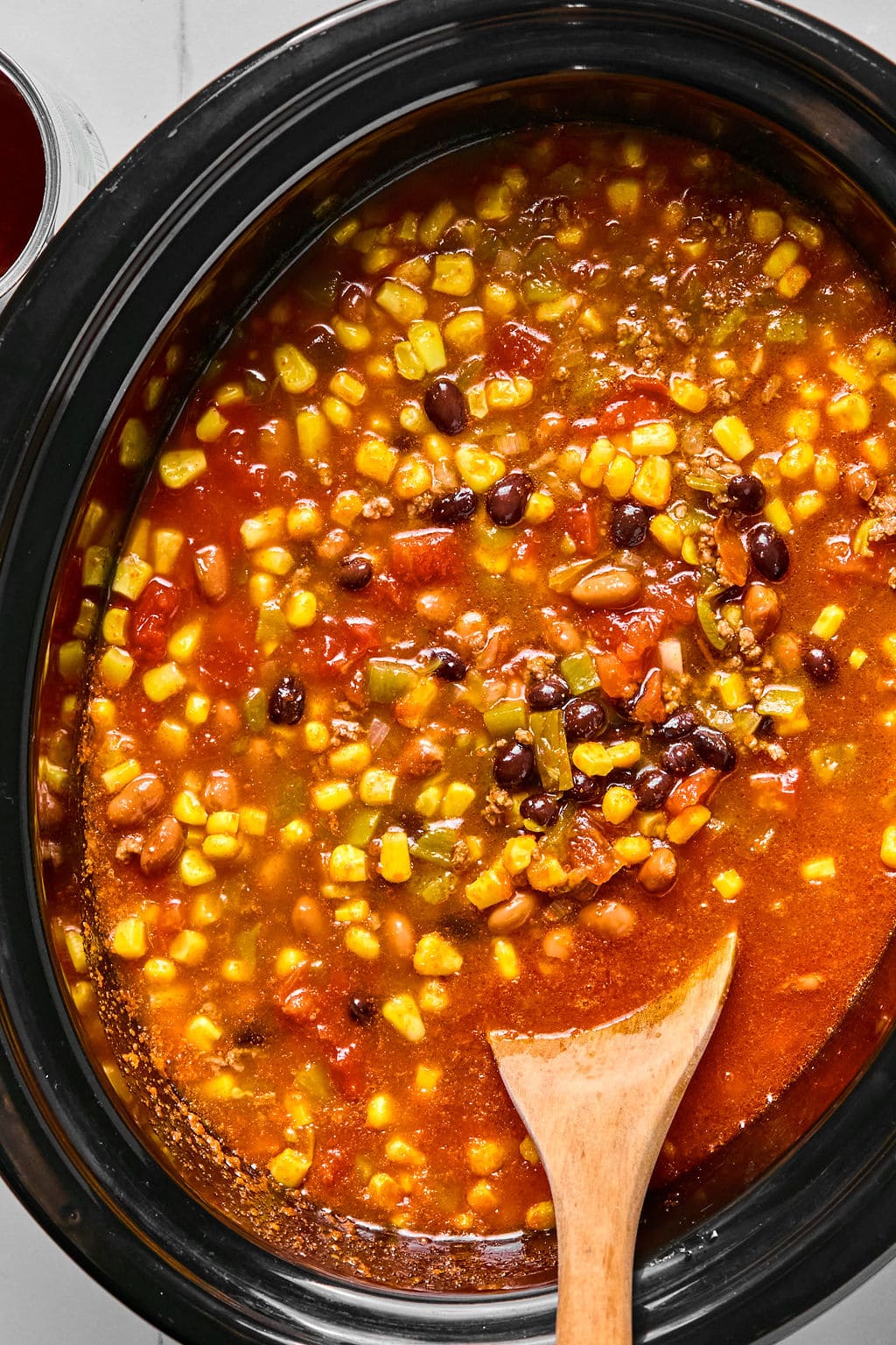 A slow cooker filled with vegetable soup containing corn, black beans, tomatoes, and other mixed vegetables. A wooden spoon rests in the soup.