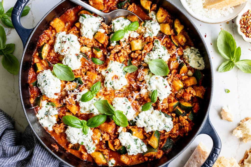 Cast iron skillet filled with a tomato-based mixture of meat, zucchini, and dollops of ricotta, garnished with fresh basil leaves. Slices of bread and basil are arranged nearby on the table.