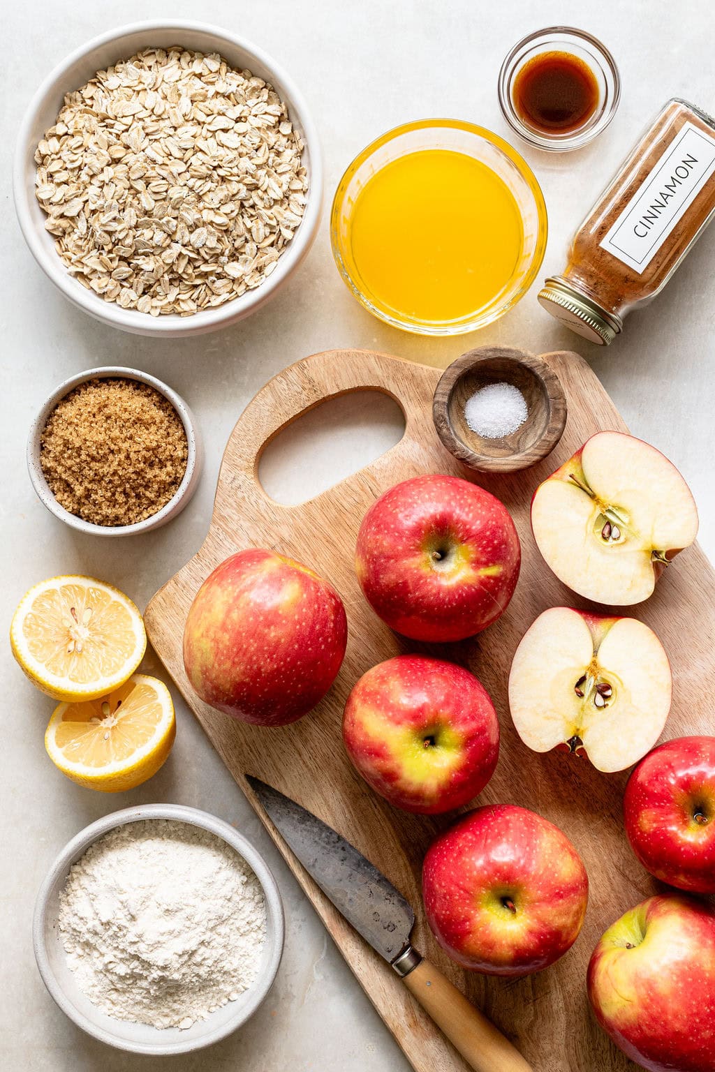 A cutting board with whole and halved apples, a knife, and bowls of oats, flour, brown sugar, orange juice, cinnamon, vanilla, salt, and lemon halves arranged on a light surface.