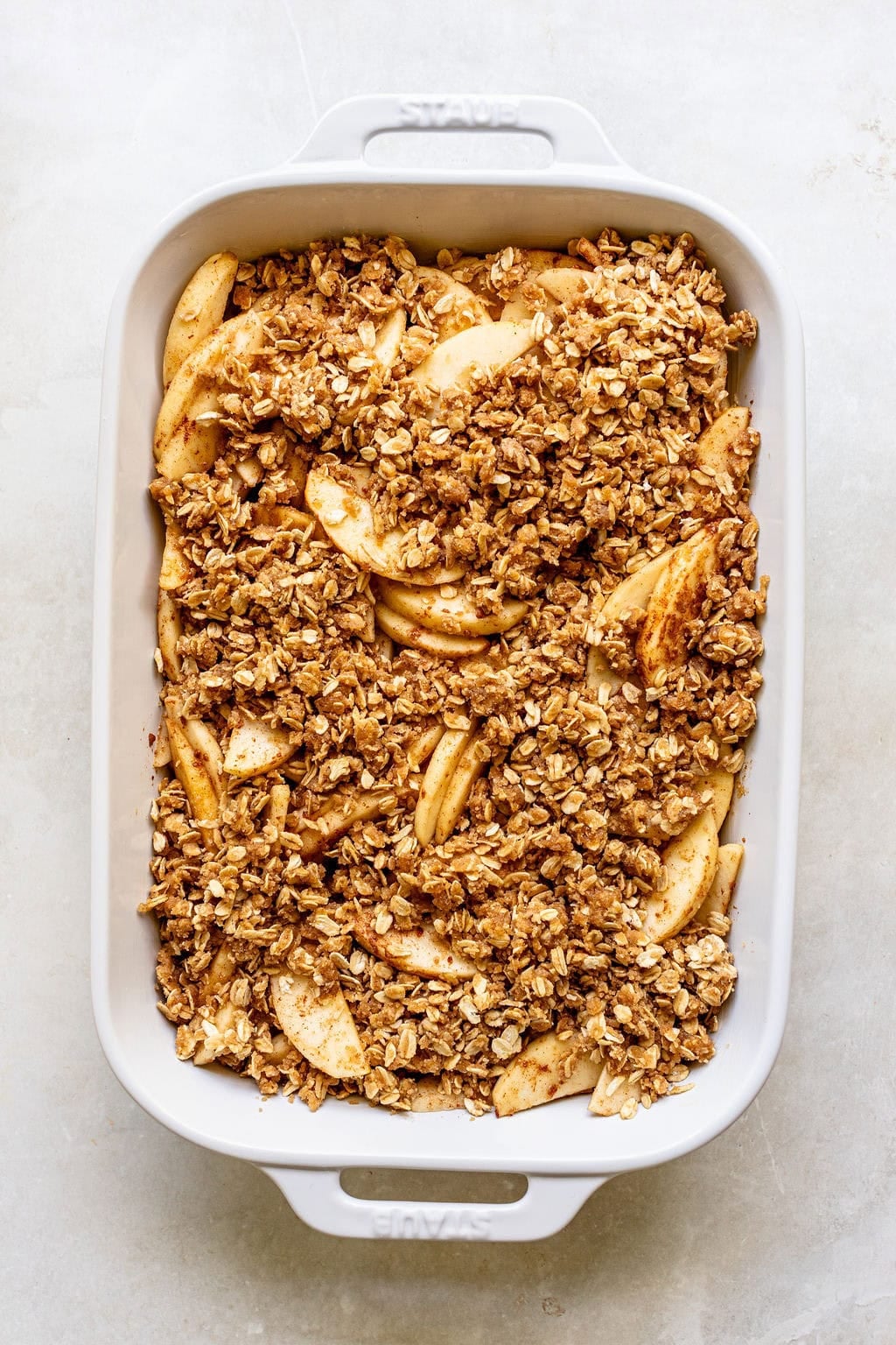 White baking dish filled with sliced apples topped with a crumbly oat mixture, ready to be baked for apple crisp.