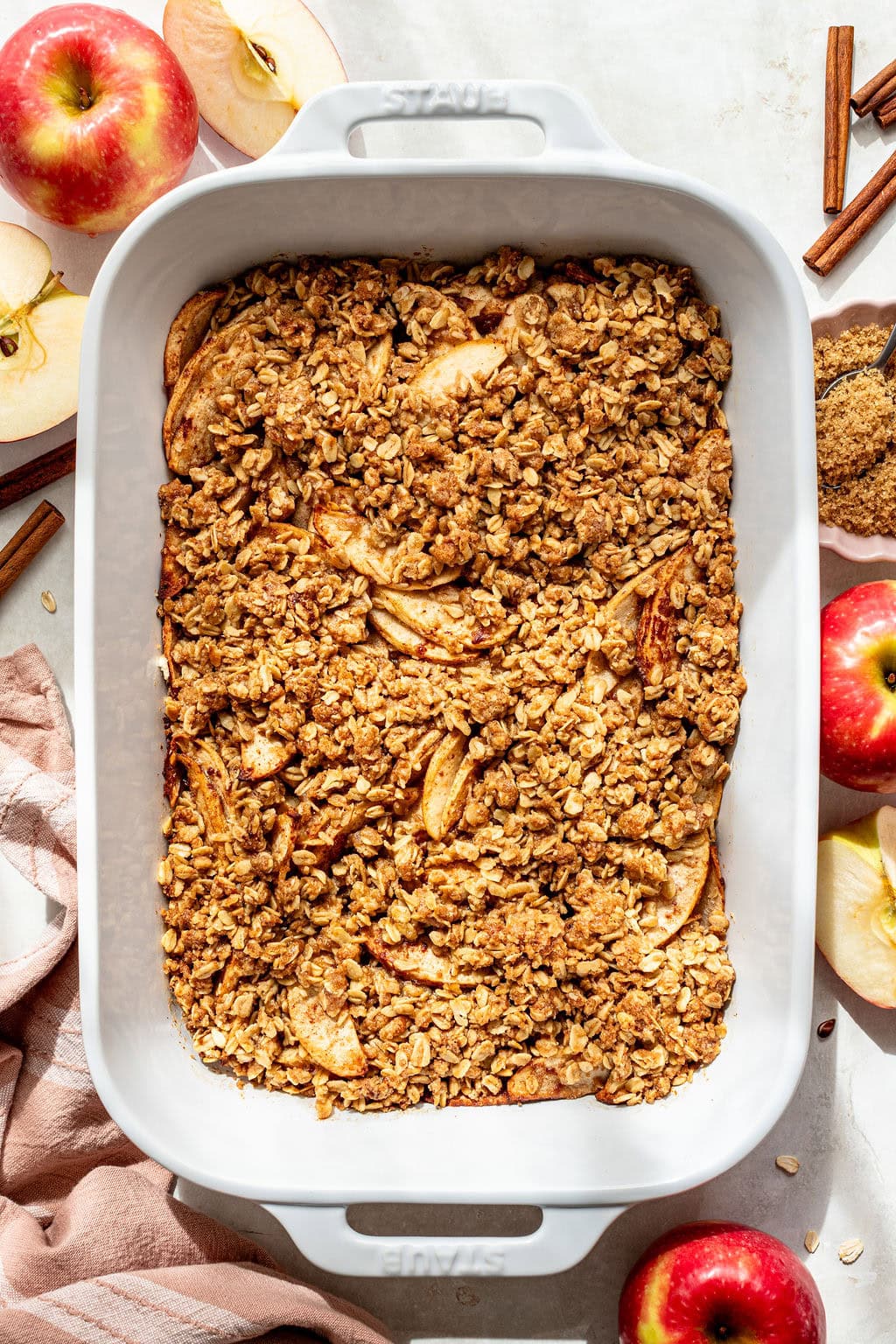 Baked apple crisp in a white dish, surrounded by fresh apples, apple slices, cinnamon sticks, and brown sugar on a light surface.
