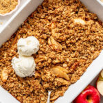 Baked apple crisp in a white dish, topped with two scoops of vanilla ice cream. Fresh apples, cinnamon sticks, and oats are arranged nearby.