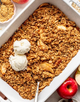 Baked apple crisp in a white dish, topped with two scoops of vanilla ice cream. Fresh apples, cinnamon sticks, and oats are arranged nearby.