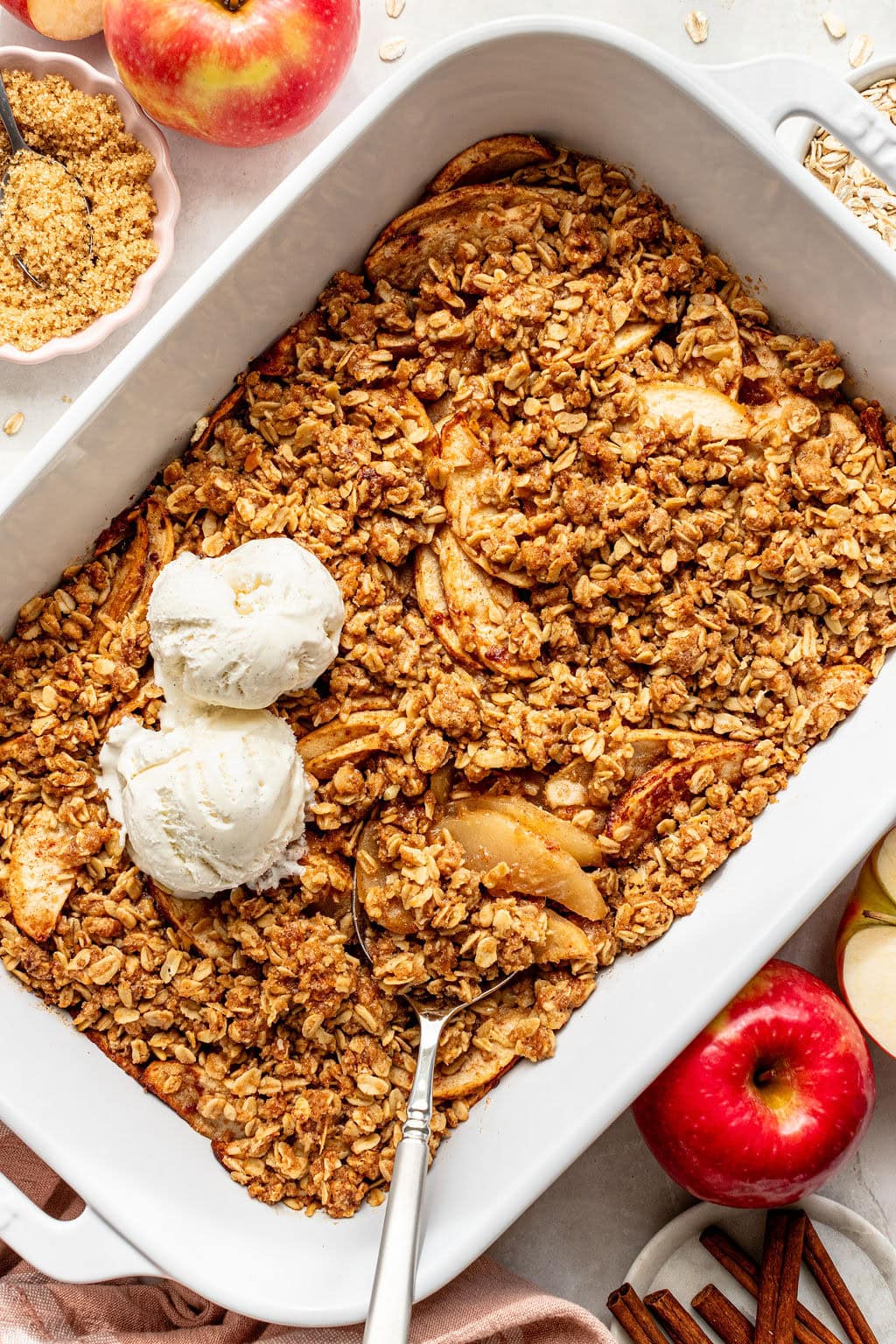 Baked apple crisp in a white dish, topped with two scoops of vanilla ice cream. Fresh apples, cinnamon sticks, and oats are arranged nearby.