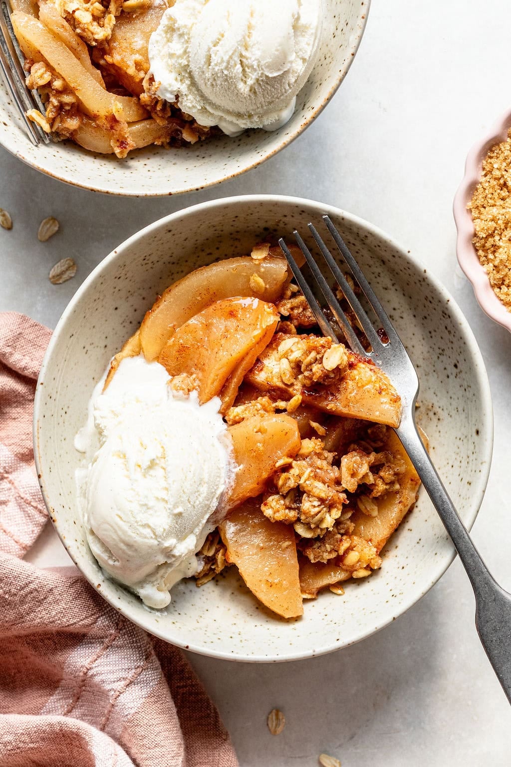 A bowl of apple crisp topped with a scoop of vanilla ice cream sits beside a fork and a pink napkin on a white surface.