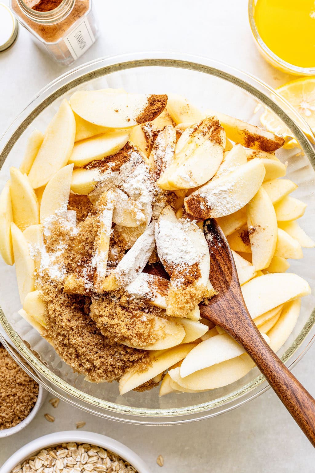Sliced apples in a glass bowl with brown sugar, flour, and spices, being mixed with a wooden spoon; ingredients and juice are visible nearby.