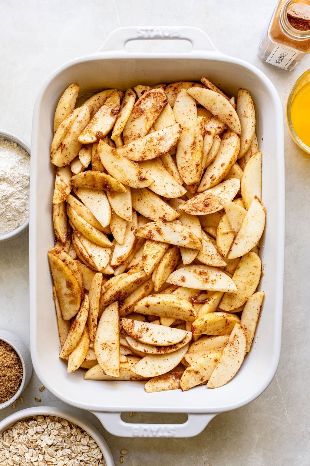 Sliced apples coated with cinnamon are arranged in a white baking dish, surrounded by bowls containing flour, brown sugar, oats, orange juice, and a jar of cinnamon.