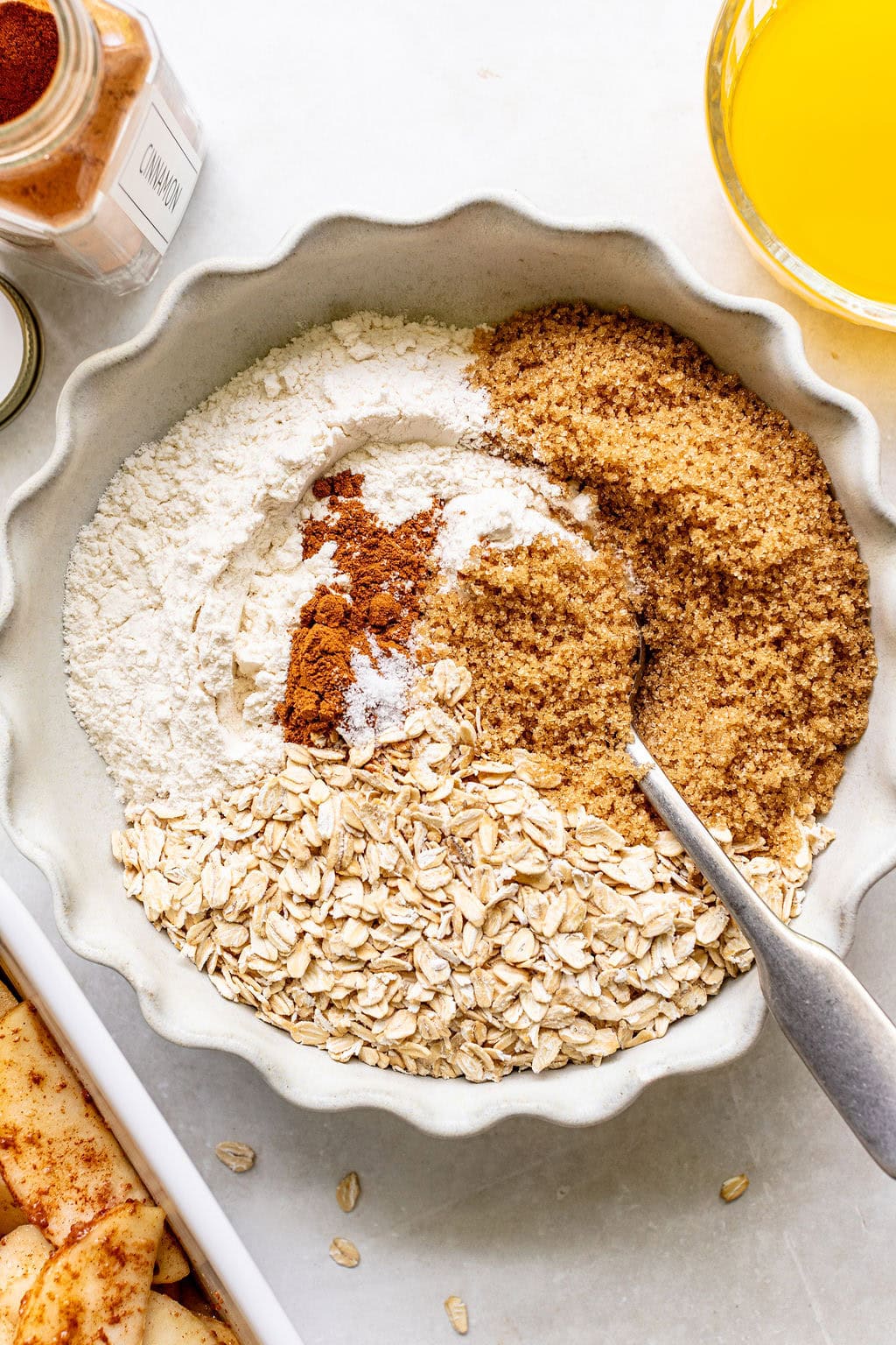 A bowl contains flour, rolled oats, brown sugar, and spices with a spoon. Nearby are a jar of cinnamon, a glass of orange liquid, and a dish with sliced apples.