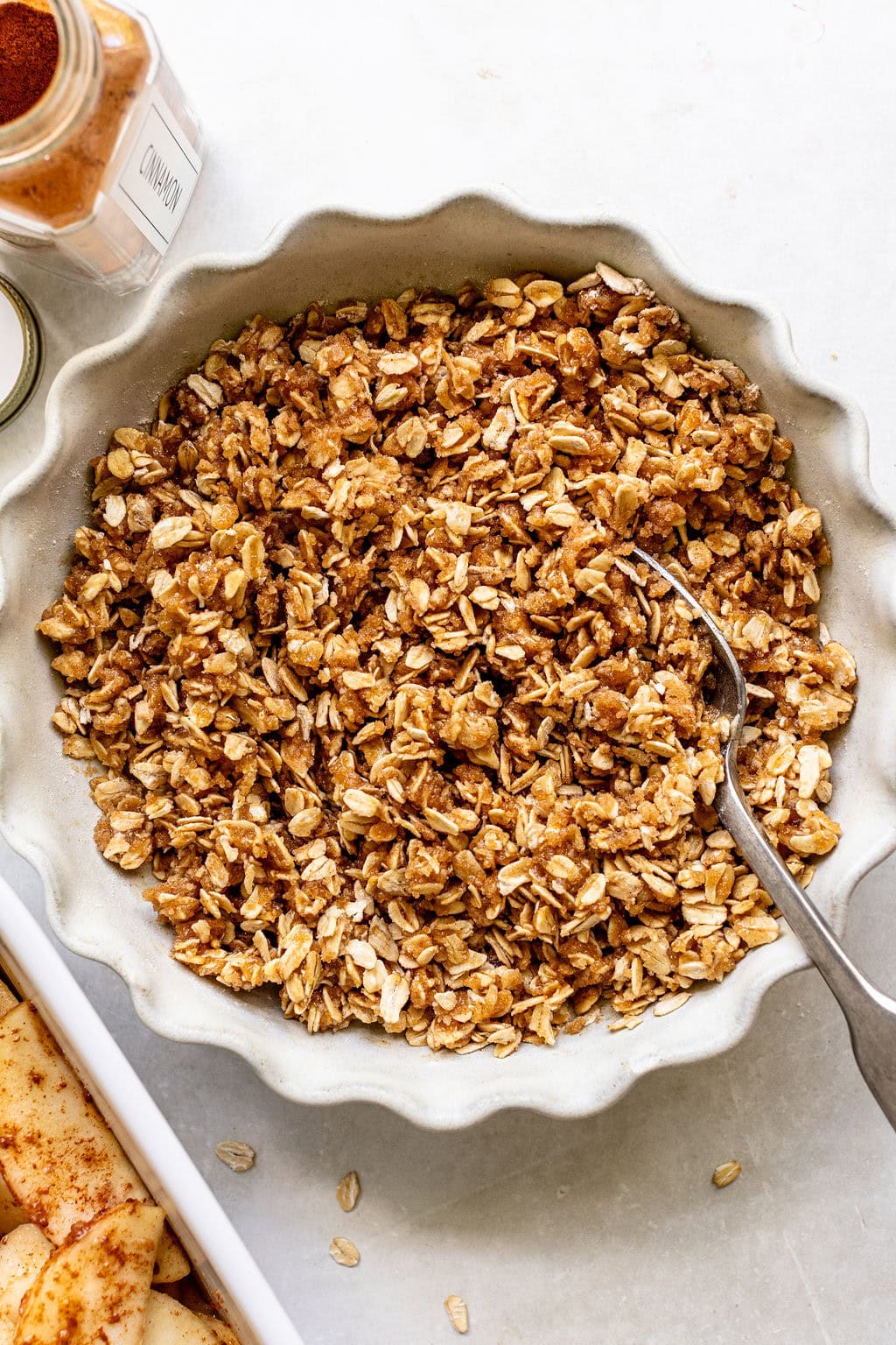 A round dish filled with oat and brown sugar crumble topping, with a spoon resting inside. A jar of cinnamon and a glimpse of sliced apples are nearby on a white surface.