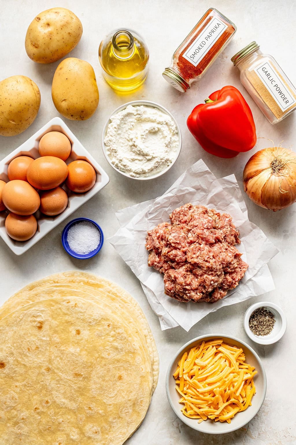 Assorted ingredients on a white surface including eggs, potatoes, ground meat, tortillas, cheese, bell pepper, onion, spices, oil, ricotta, salt, and pepper.