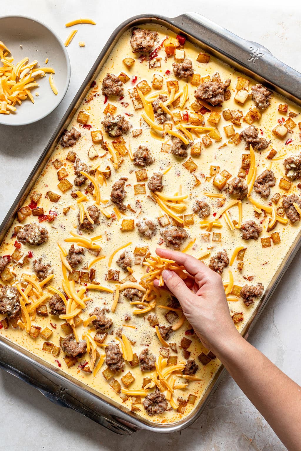 A hand sprinkles shredded cheese over a breakfast casserole in a metal baking pan, topped with ground meet and diced vegetables. A small bowl of extra cheese sits nearby.