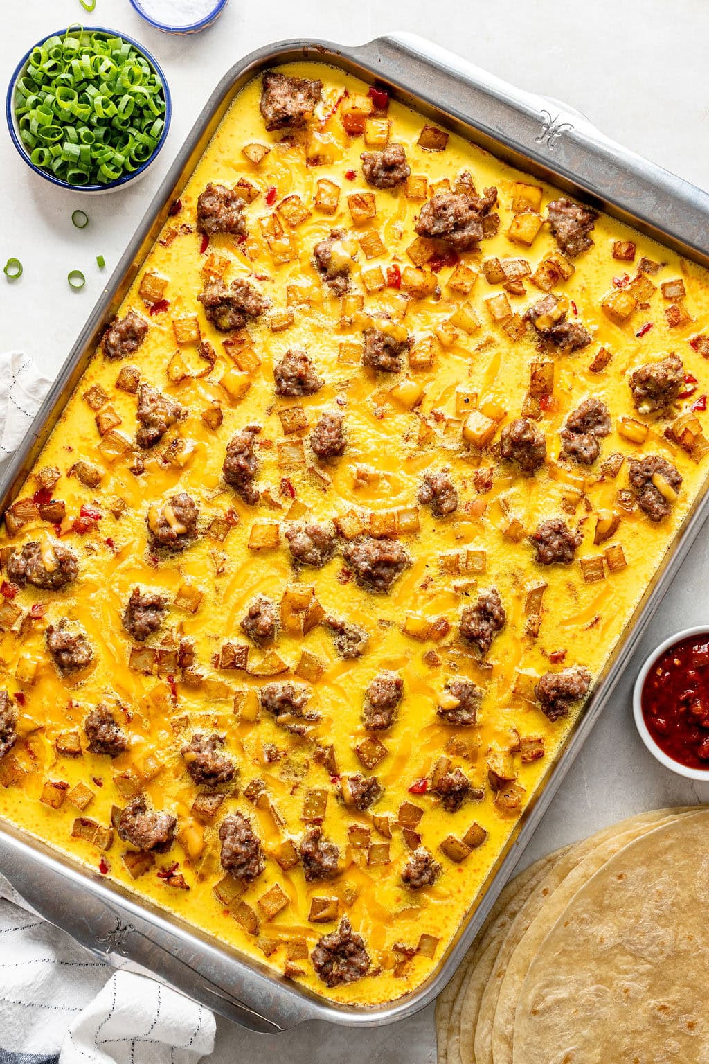 Sheet pan filled with baked egg casserole topped with sausage, diced potatoes, and cheese. Bowls of sliced green onions and salsa, as well as tortillas, are arranged nearby on the table.