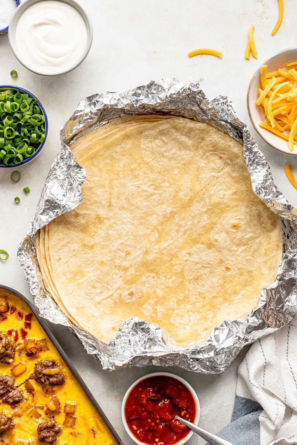 A stack of tortillas in foil surrounded by bowls of sour cream, shredded cheese, chopped green onions, salsa, and a sheet pan with meat and cheese.