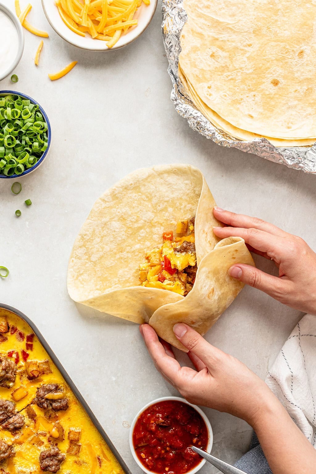 Hands folding a tortilla filled with cooked meat, eggs, and potatoes; bowls of salsa, shredded cheese, green onions, and a stack of tortillas are nearby on a light surface.
