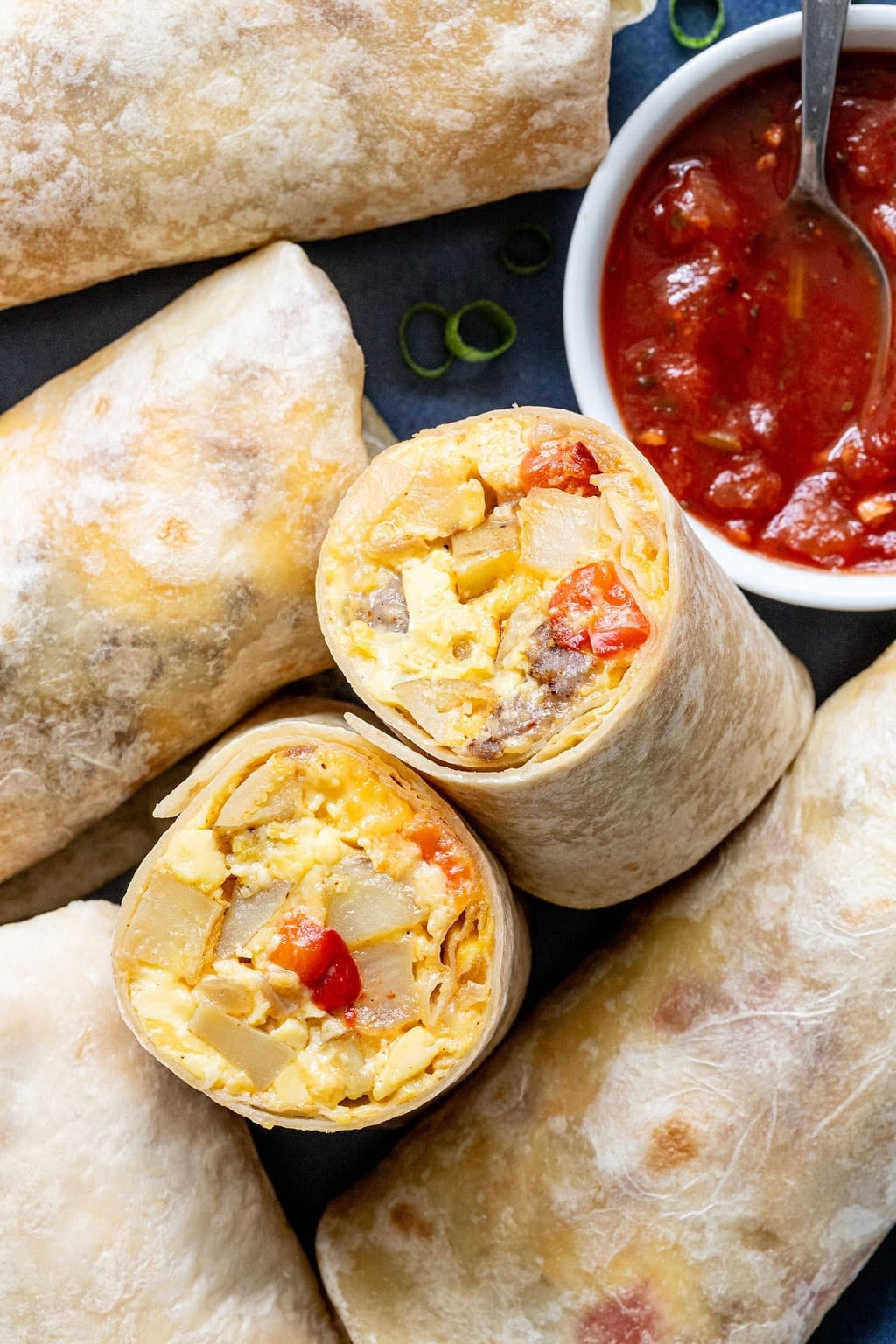 Breakfast burritos filled with eggs, potatoes, sausage, and cheese are shown on a plate next to a bowl of chunky tomato salsa with a spoon.