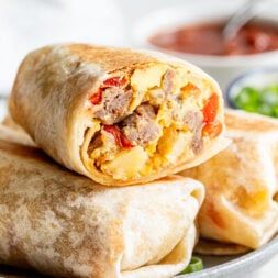 A close-up of a breakfast burrito cut in half, showing scrambled eggs, sausage, and cheese, stacked on a plate with a bowl of salsa in the background.
