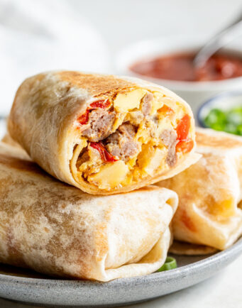 A close-up of a breakfast burrito cut in half, showing scrambled eggs, sausage, and cheese, stacked on a plate with a bowl of salsa in the background.