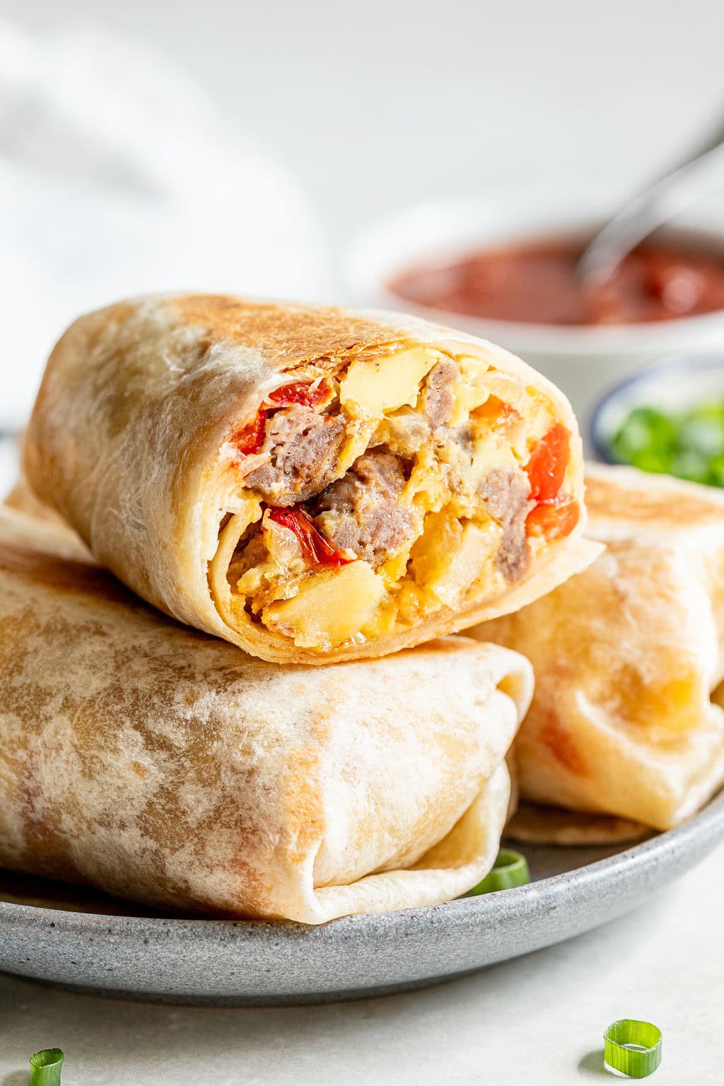 A close-up of a breakfast burrito cut in half, showing scrambled eggs, sausage, and cheese, stacked on a plate with a bowl of salsa in the background.