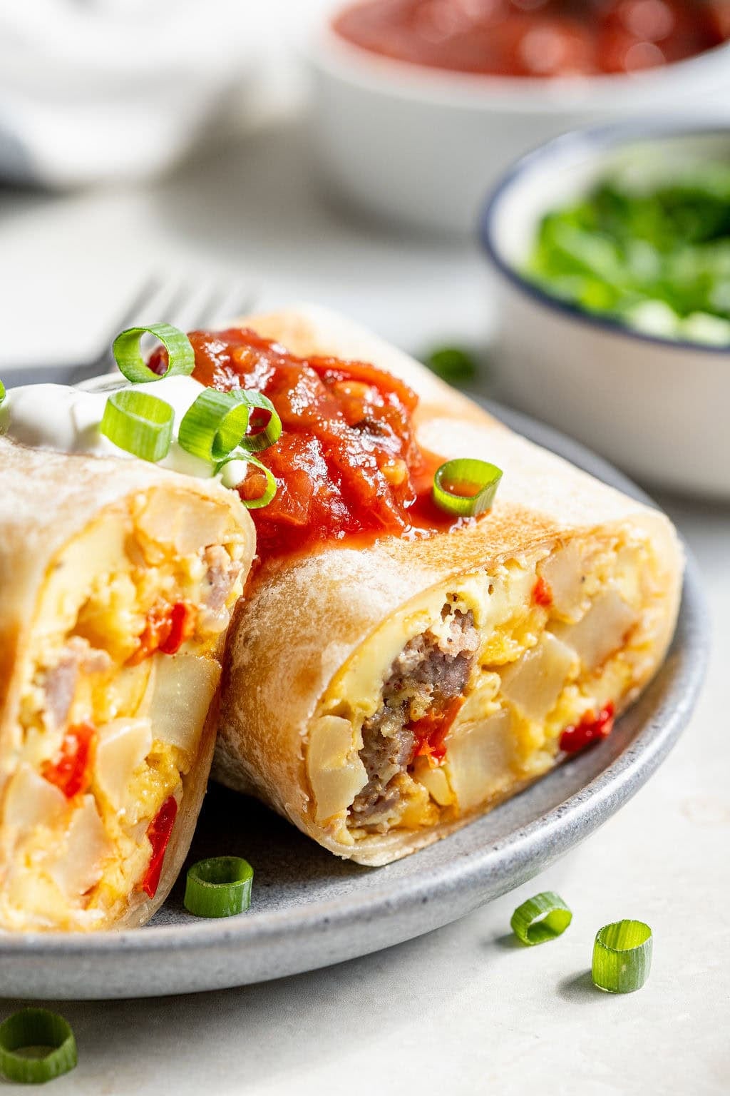 A breakfast burrito cut in half on a plate, filled with eggs, sausage, potatoes, and red peppers, topped with salsa and garnished with chopped green onions.