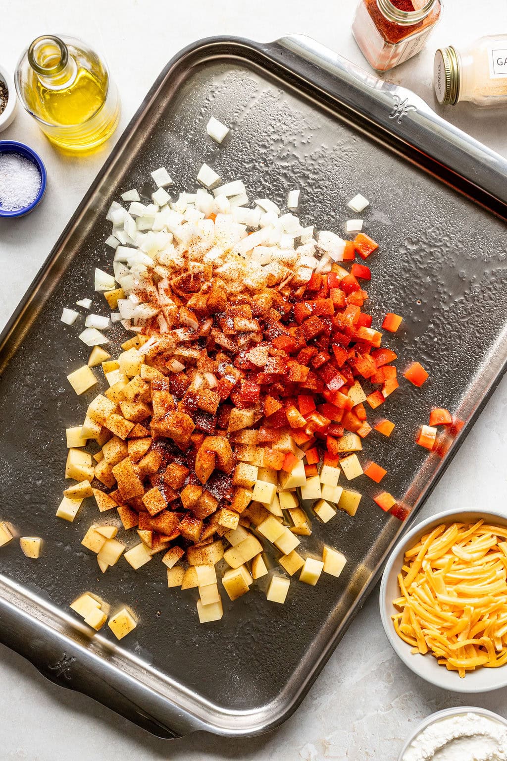 A baking sheet with chopped potatoes, onions, and red bell peppers sprinkled with spices, surrounded by bowls of shredded cheese, flour, oil, and seasonings.