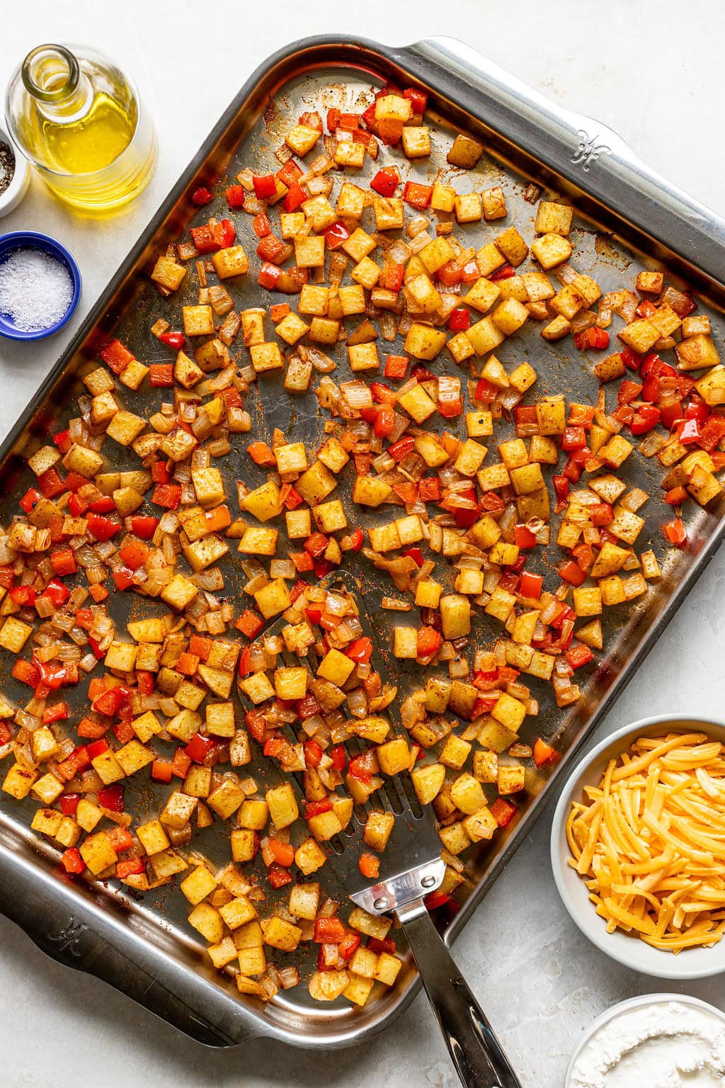 A baking sheet with roasted diced potatoes and red bell peppers, surrounded by bowls of shredded cheese, olive oil, and salt.