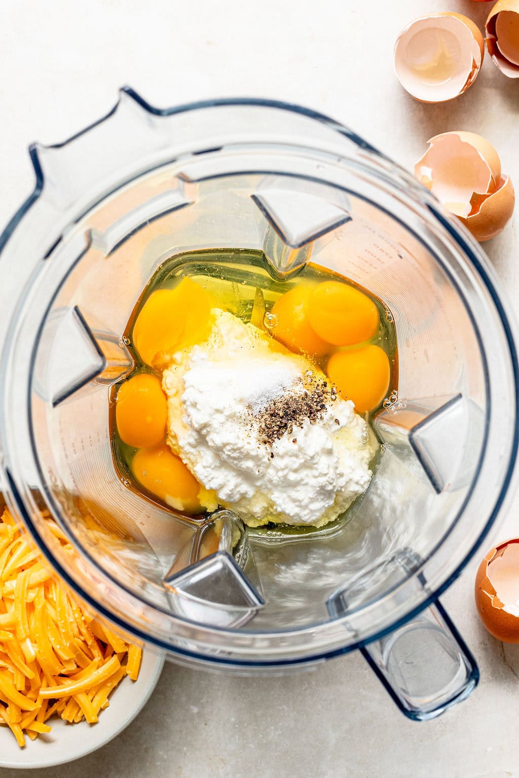 A blender containing eggs, ricotta cheese, and pepper, with a bowl of shredded cheddar and cracked eggshells nearby on a white surface.