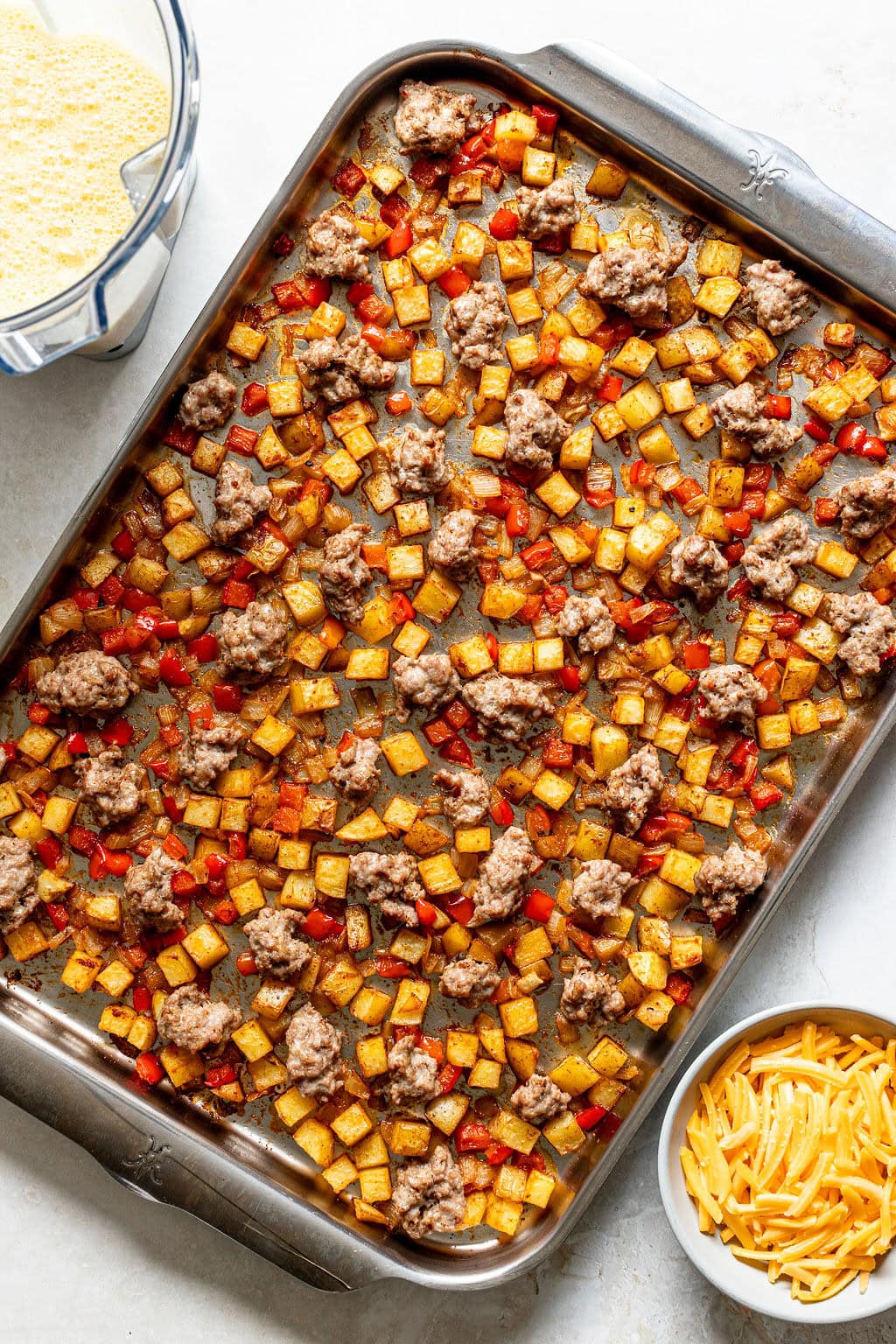 A baking sheet with cooked diced potatoes, red bell peppers, and crumbled ground beef. A bowl of shredded cheese and a blender of beaten eggs are placed nearby.
