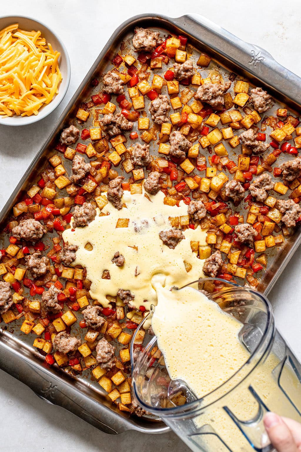 A baking sheet with diced potatoes, red peppers, ground meat, and egg mixture being poured on top; a bowl of shredded cheese is nearby.