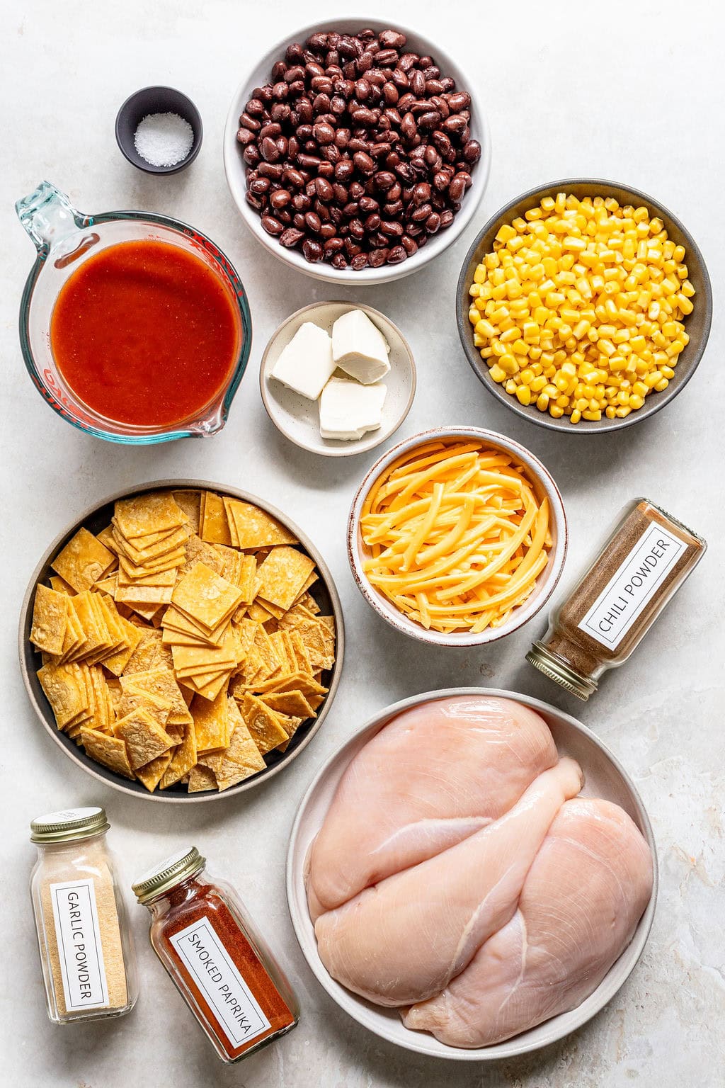 Ingredients for enchiladas, including raw chicken breasts, black beans, corn, cheese, tortilla chips, tomato sauce, cream cheese, and spices, arranged in bowls on a white surface.