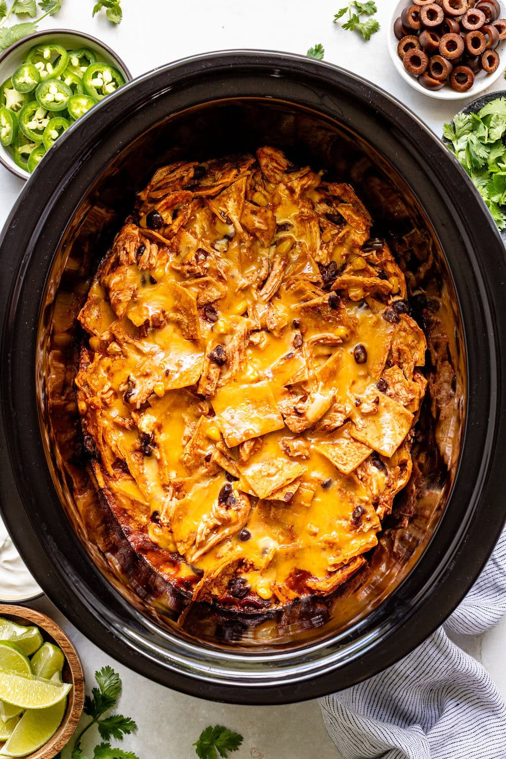 A slow cooker filled with cheesy chicken enchilada casserole, surrounded by bowls of jalapeños, olives, lime wedges, sour cream, and fresh cilantro.