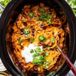 Chicken enchilada casserole in a slow cooker, topped with sliced olives, jalapeños, sour cream, cheese, and cilantro. Bowls of limes, jalapeños, and olives are nearby.