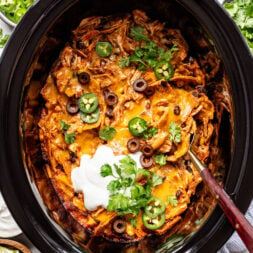 Chicken enchilada casserole in a slow cooker, topped with sliced olives, jalapeños, sour cream, cheese, and cilantro. Bowls of limes, jalapeños, and olives are nearby.