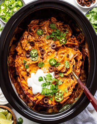 Chicken enchilada casserole in a slow cooker, topped with sliced olives, jalapeños, sour cream, cheese, and cilantro. Bowls of limes, jalapeños, and olives are nearby.