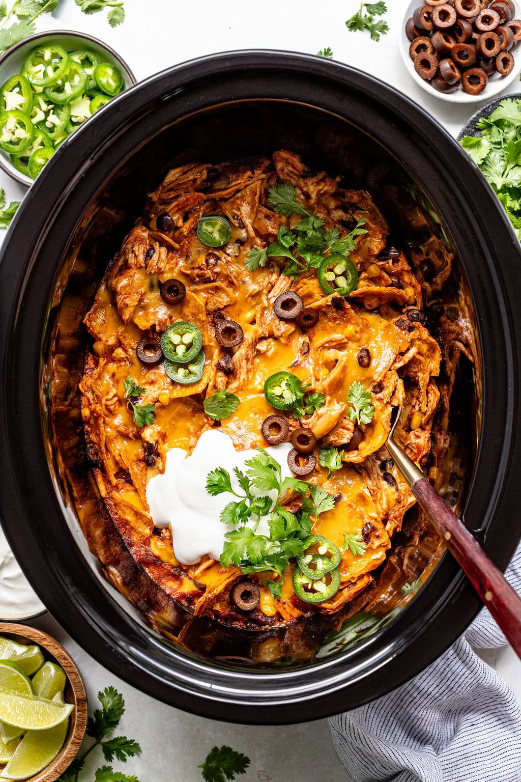 Chicken enchilada casserole in a slow cooker, topped with sliced olives, jalapeños, sour cream, cheese, and cilantro. Bowls of limes, jalapeños, and olives are nearby.