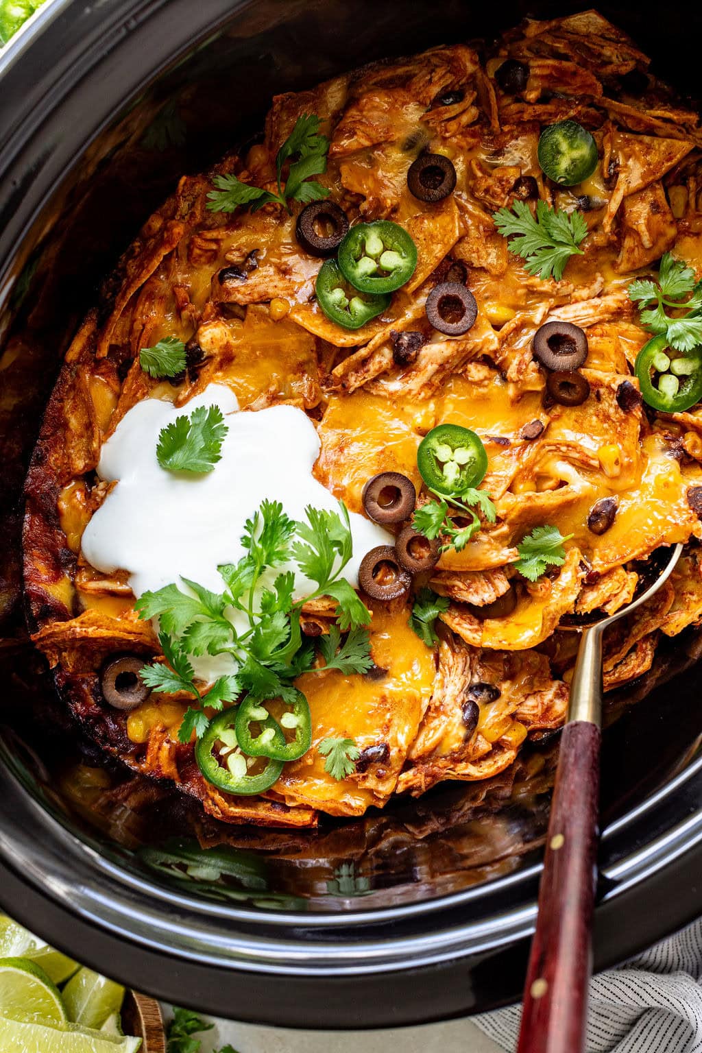 A slow cooker filled with cheesy chicken enchilada casserole, topped with sliced jalapeños, black olives, cilantro, and a dollop of sour cream.